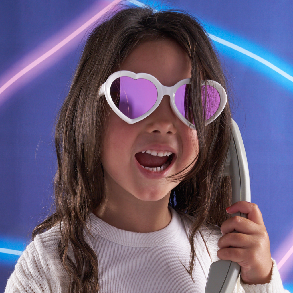 A young girl with long brown hair wearing white heart-shaped sunglasses with purple mirrored lenses, holding a gray toy phone against a blue background with neon light streaks