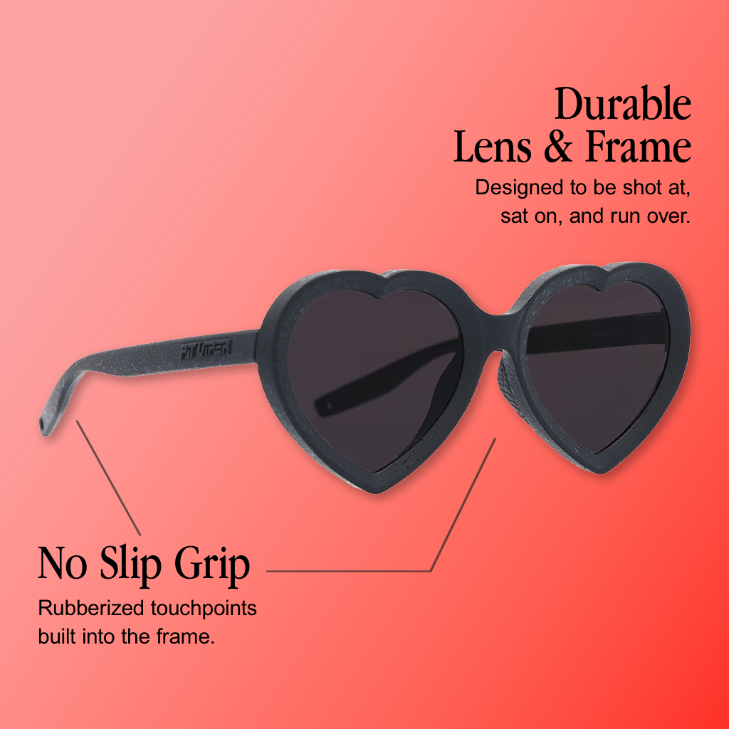 Infographic for Pit Viper Admirer sunglasses showing black heart-shaped frame sunglasses with dark smoke lenses and 'PIT VIPER' embossed on the temple arm, angled against a pink-to-coral gradient background, with 2 callouts: 'Durable Lens & Frame - Designed to be shot at, sat on, and run over.', 'No Slip Grip - Rubberized touchpoints built into the frame.'