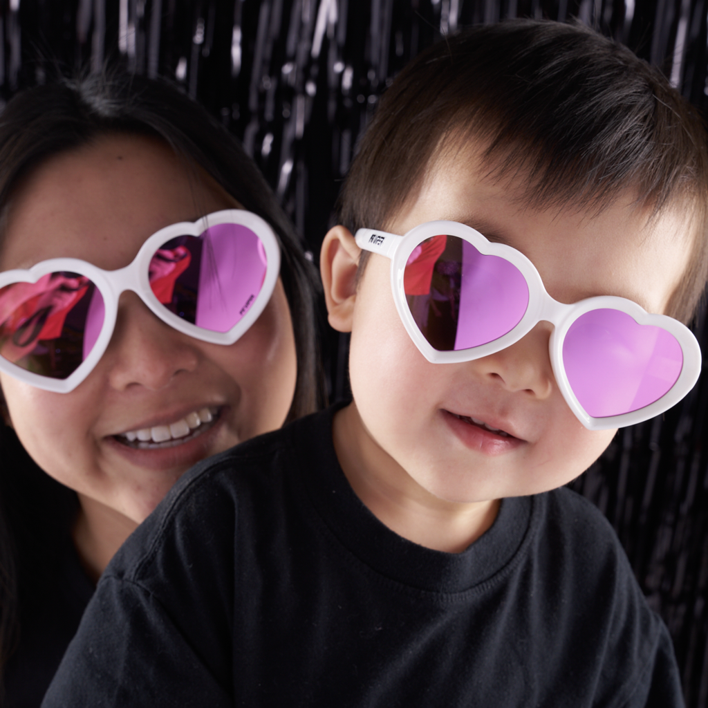 A close-up lifestyle shot of a woman and a young child wearing white heart-shaped sunglasses with pink-tinted lenses against a black tinsel backdrop.