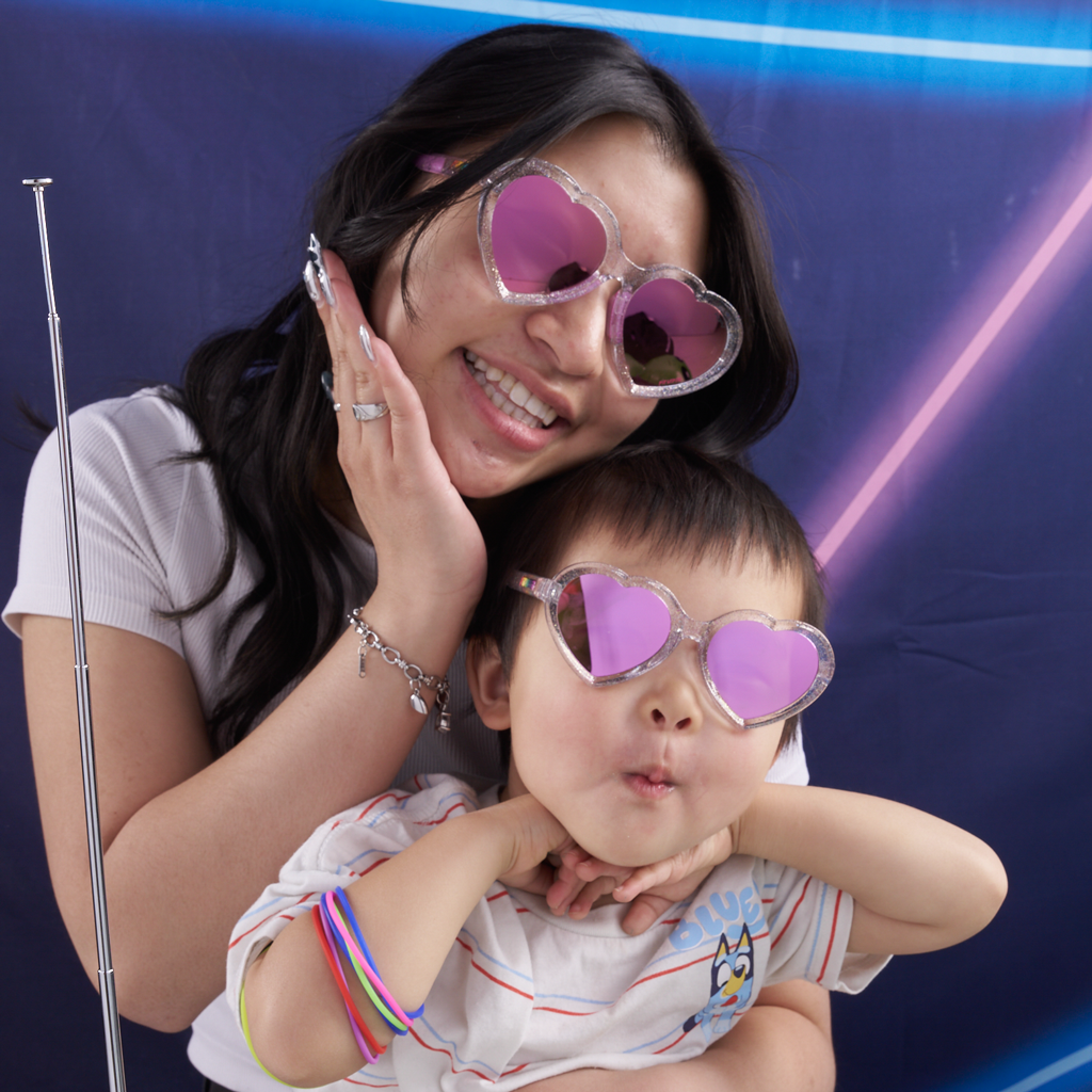 A smiling woman and a young child wearing heart-shaped pink sunglasses pose together in front of a blue photo booth backdrop with neon light effects.