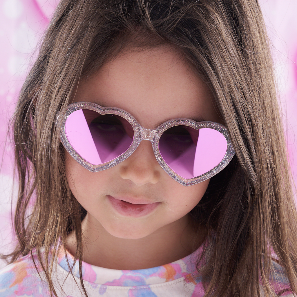 A close-up of a young girl with long brown hair wearing heart-shaped sunglasses with glittery silver frames and pink mirrored lenses, set against a soft pink background