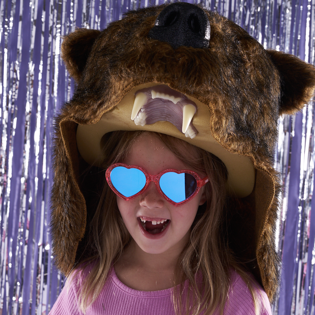 A young girl wearing a brown bear costume hat with floppy ears, faux fur, a black snout, and large white teeth, paired with red glitter heart-shaped sunglasses with blue mirrored lenses, posed in front of a purple tinsel backdrop.