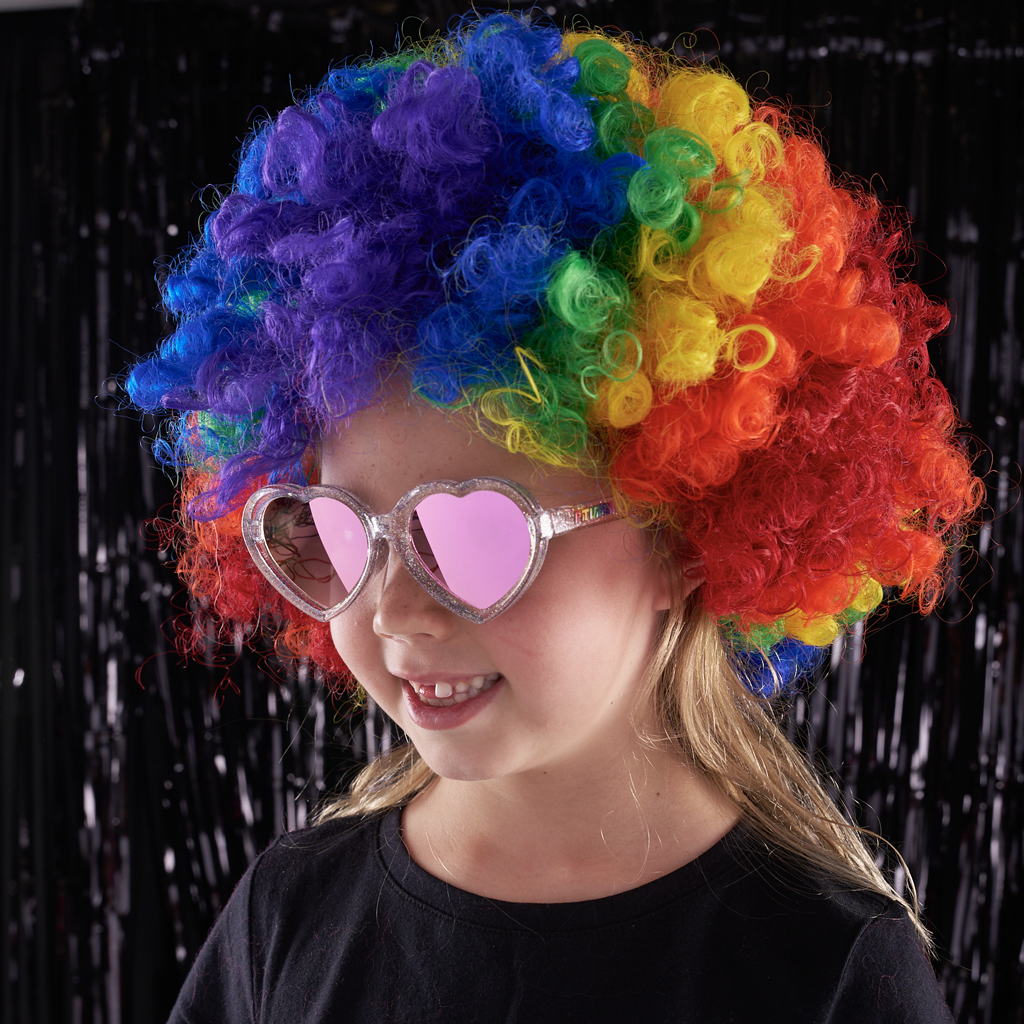 A young girl wearing a vibrant rainbow-colored curly wig and heart-shaped pink sunglasses, posing against a black tinsel curtain background.