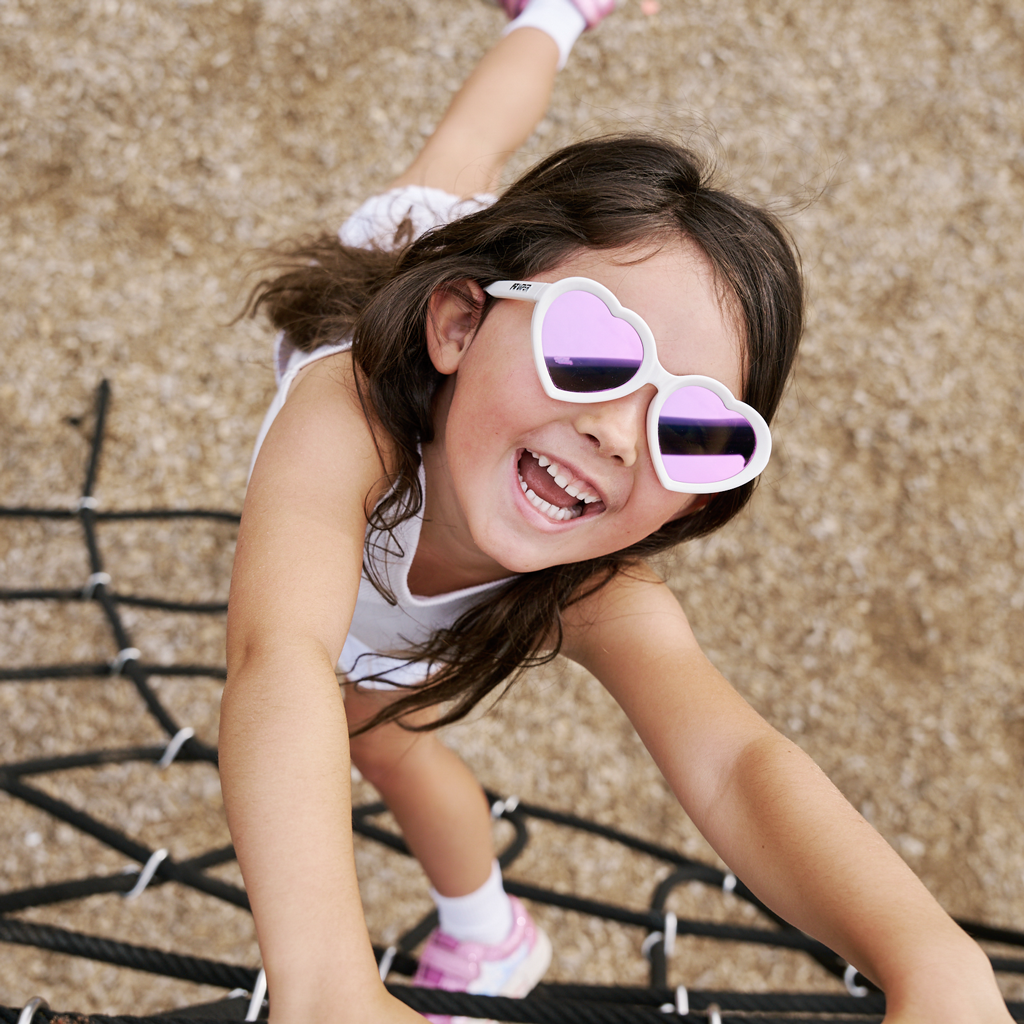 A young girl smiling and crawling on a playground rope net, wearing white heart-shaped sunglasses with purple mirrored lenses