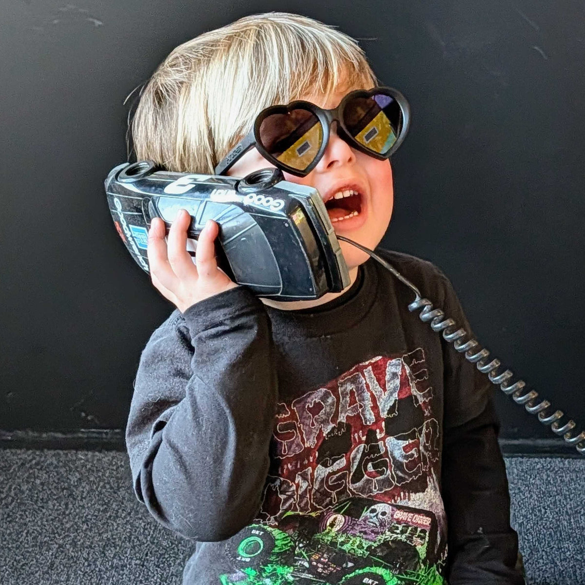 A young child sitting on a dark carpeted floor, holding a toy walkie-talkie to their ear as if talking on a real phone. The child is wearing round black sunglasses, a dark gray graphic long-sleeve shirt with green monster truck graphics, and blue jeans.