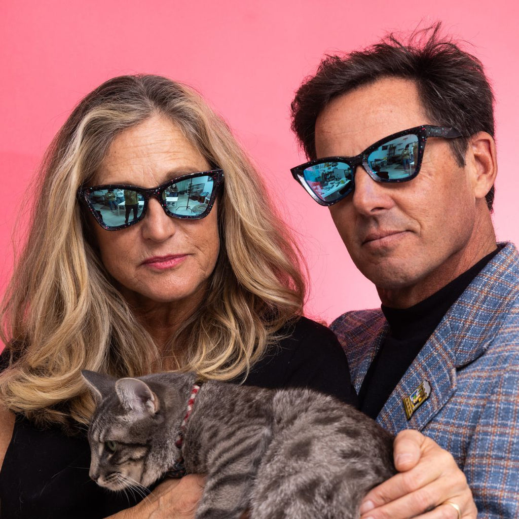 A woman and a man wear The Midnight Clawdia with a blue lens from Pit Viper while holding a cat